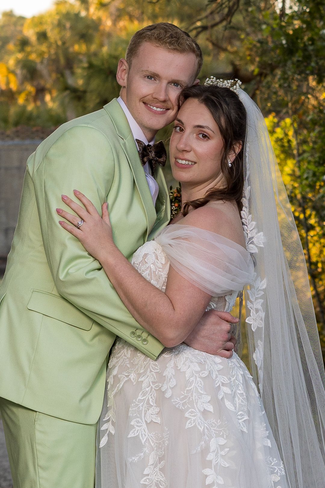 A smiling bride in a white lace gown and veil embraces a groom in a light green suit outdoors.