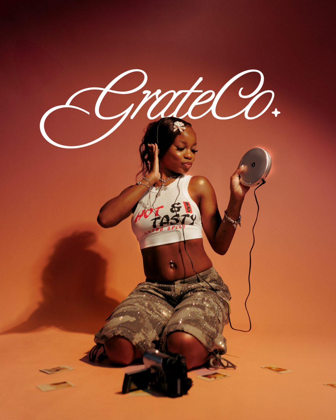Young woman kneeling in studio wearing crop top and cargo skirt, holding CD player with headphones, GrateCo branding overhead.