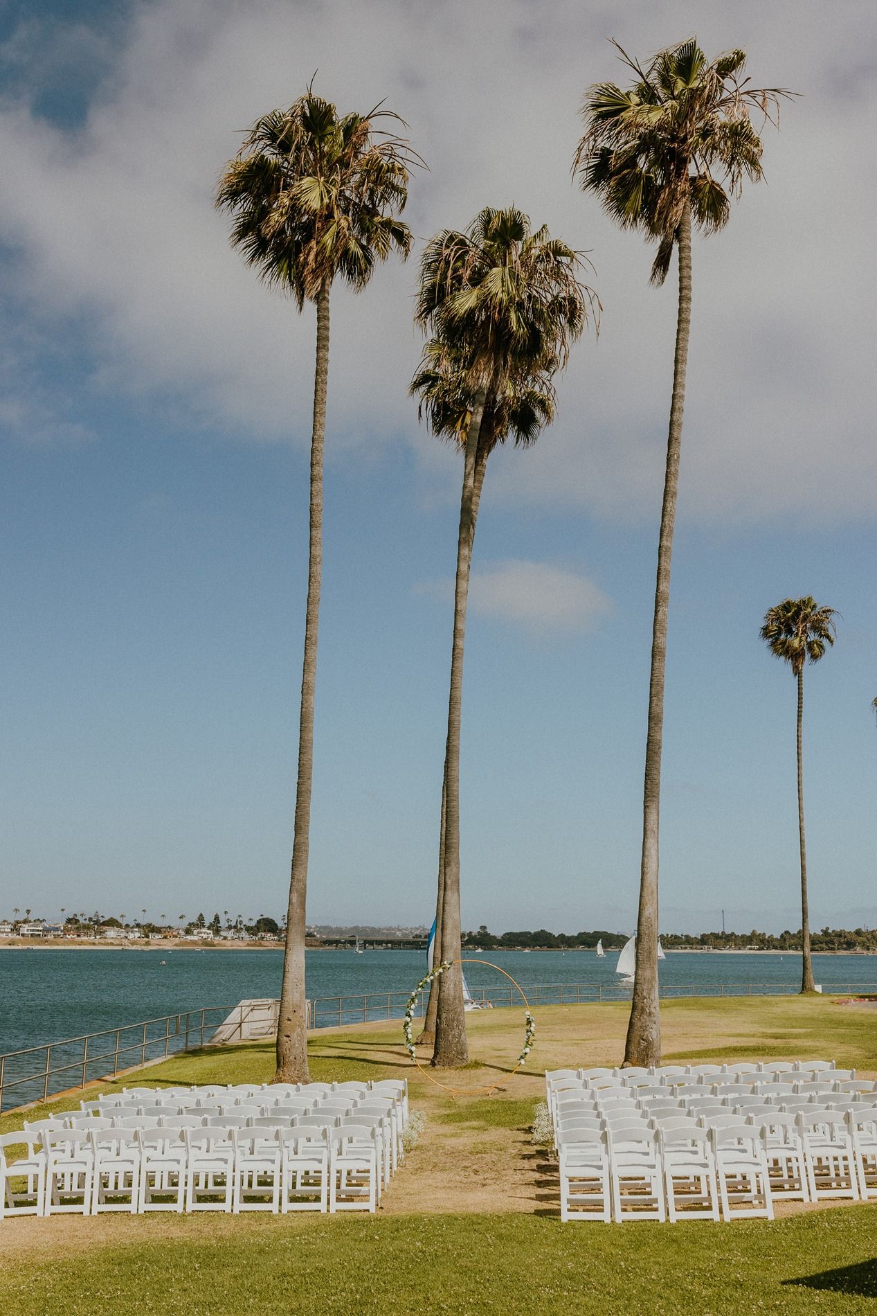 Outdoor wedding ceremony under tall palm trees by the waterfront on a sunny day with guests seated in white chairs at Tower Beach Club.