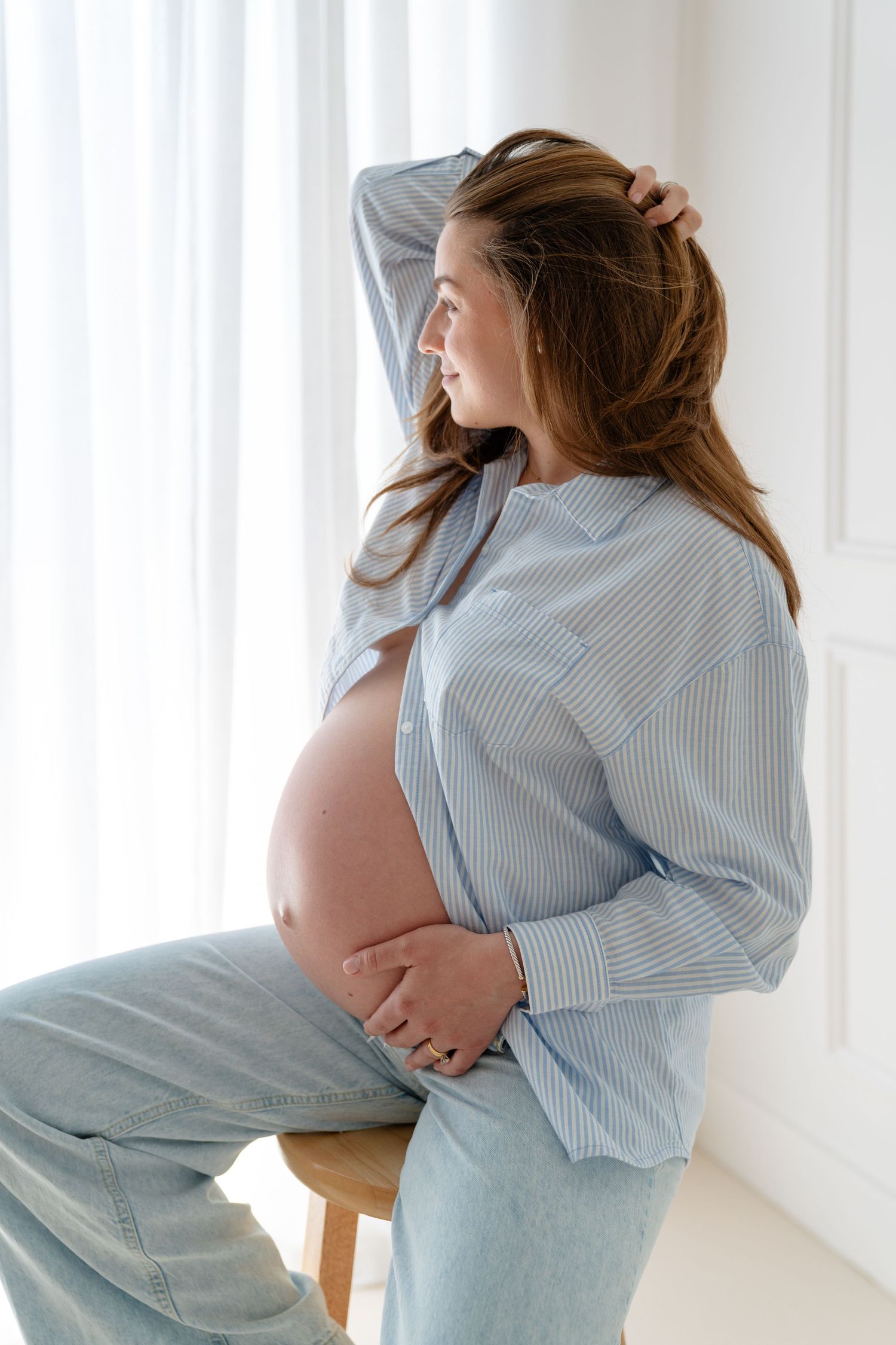 Smiling pregnant woman in blue striped shirt and jeans sitting on stool, cradling baby bump near bright window.