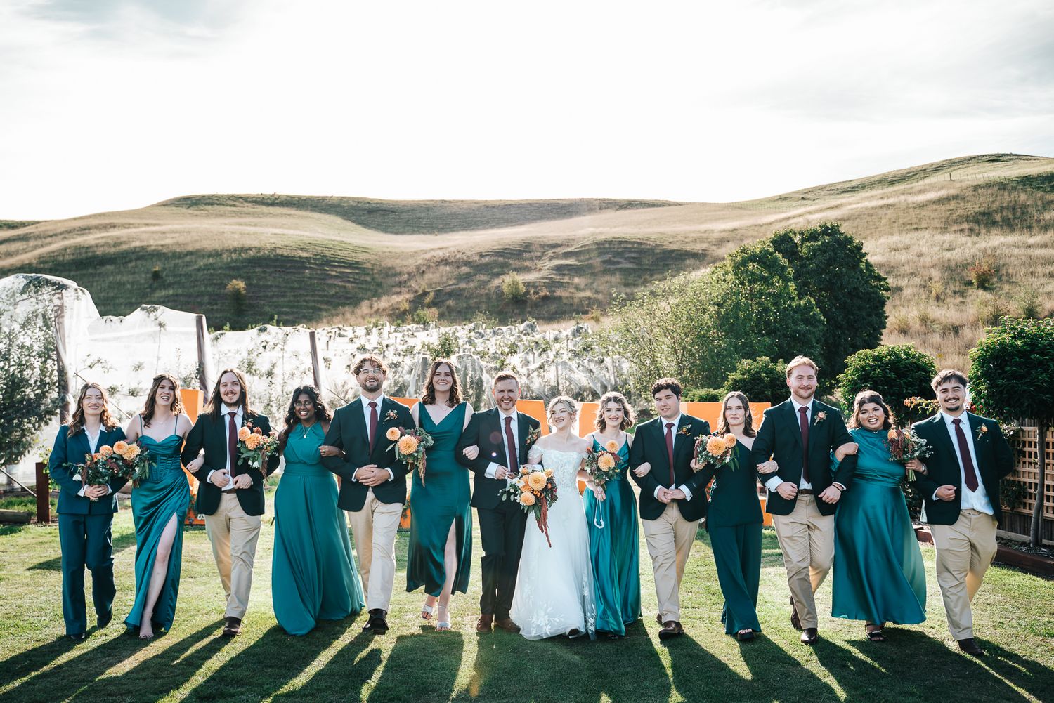 Wedding party in teal dresses and navy suits posing outdoors on green lawn with white cliffs and hills in western Melbourne.