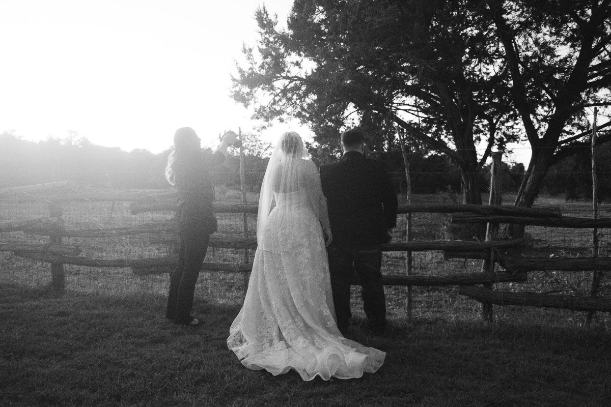 Black and white photo of a bride in a lace gown and veil standing at a rustic fence at sunset.