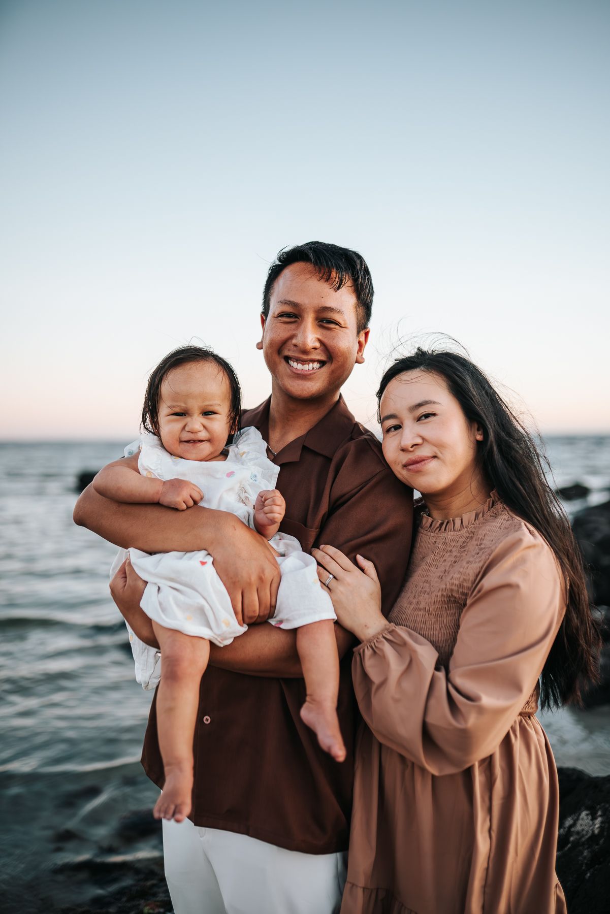 Happy family of three posing at sunset beach, parents holding baby in white dress taken during a Melbourne family photo session in Altona.