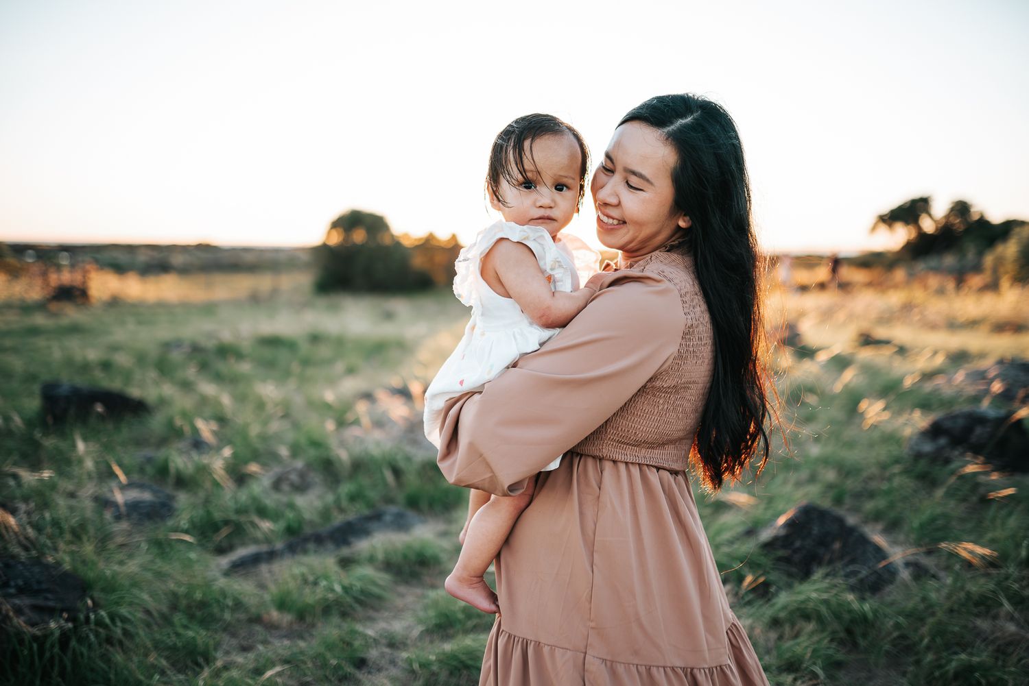 A mother tenderly holds her baby in a sunlit field at golden hour, both wearing warm-toned outfits taken by a Melbourne family photographer.