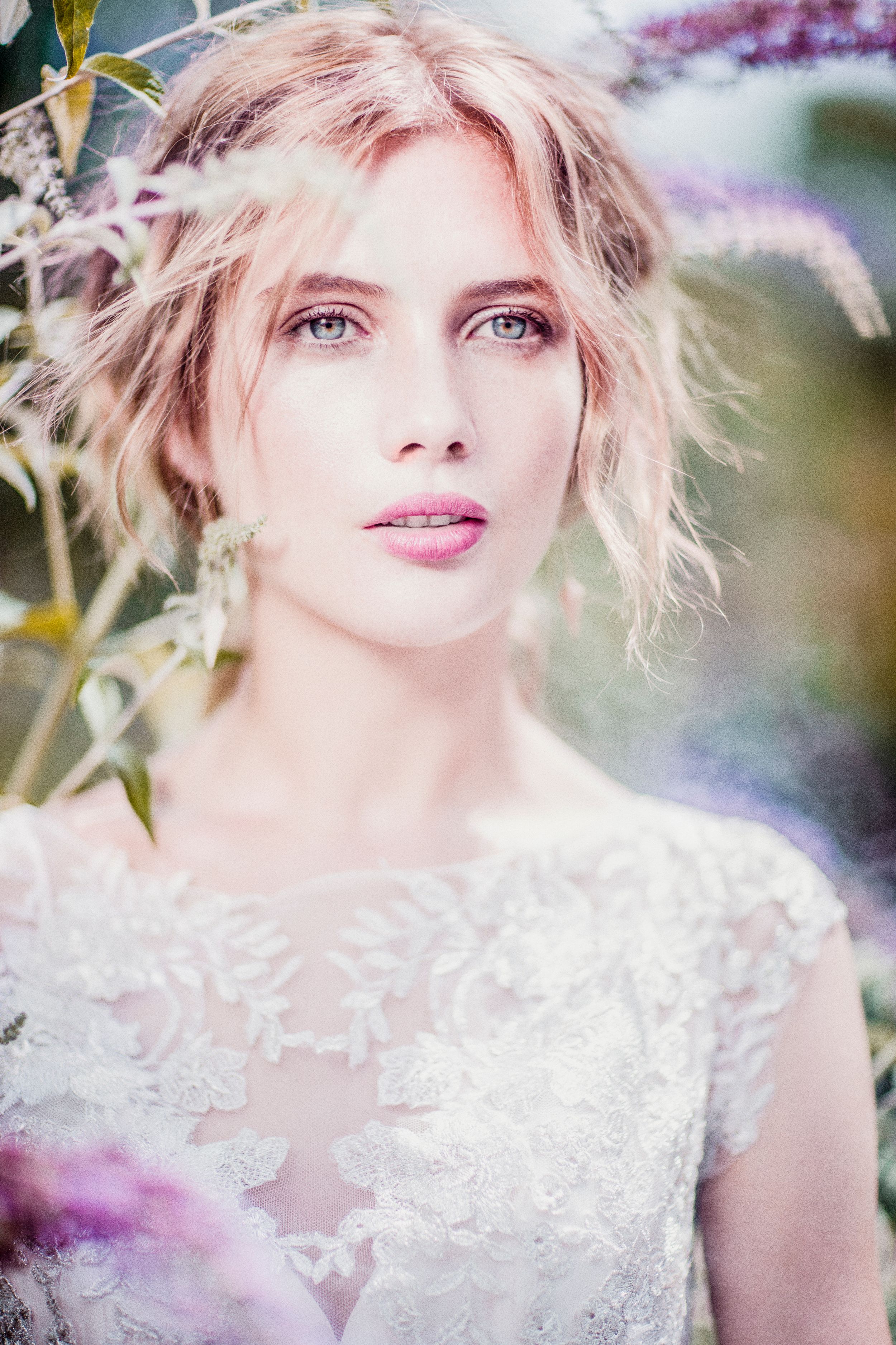 A beautiful bride in white lace dress surrounded by purple flowers with soft romantic bokeh background.