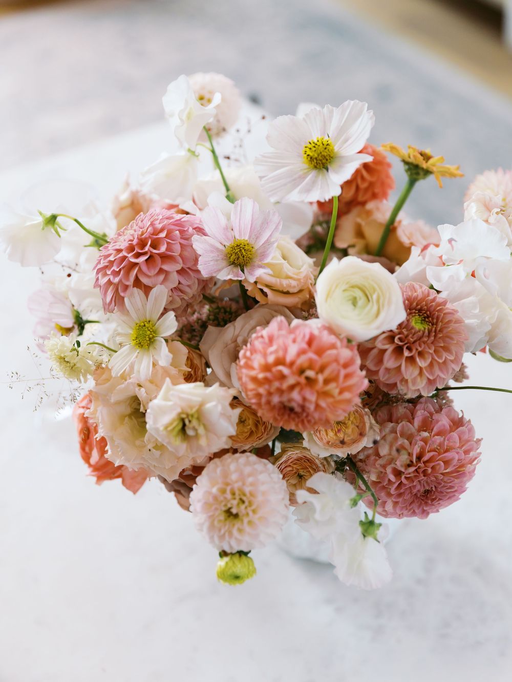 A lush bouquet of pink dahlias, white cosmos, and blush ranunculus arranged in a white vase on a marble surface.