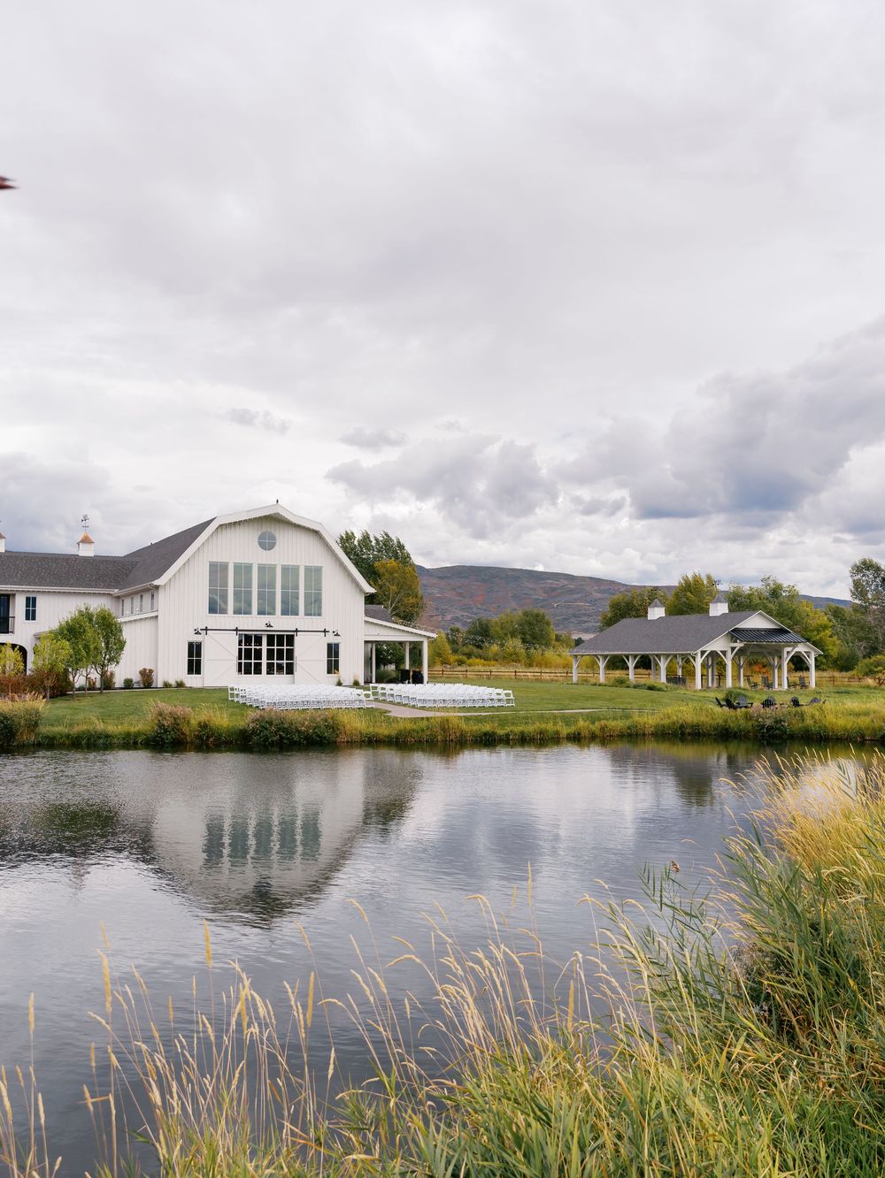 White barn venue reflects in a serene pond with mountain views and a pergola on an overcast day.
