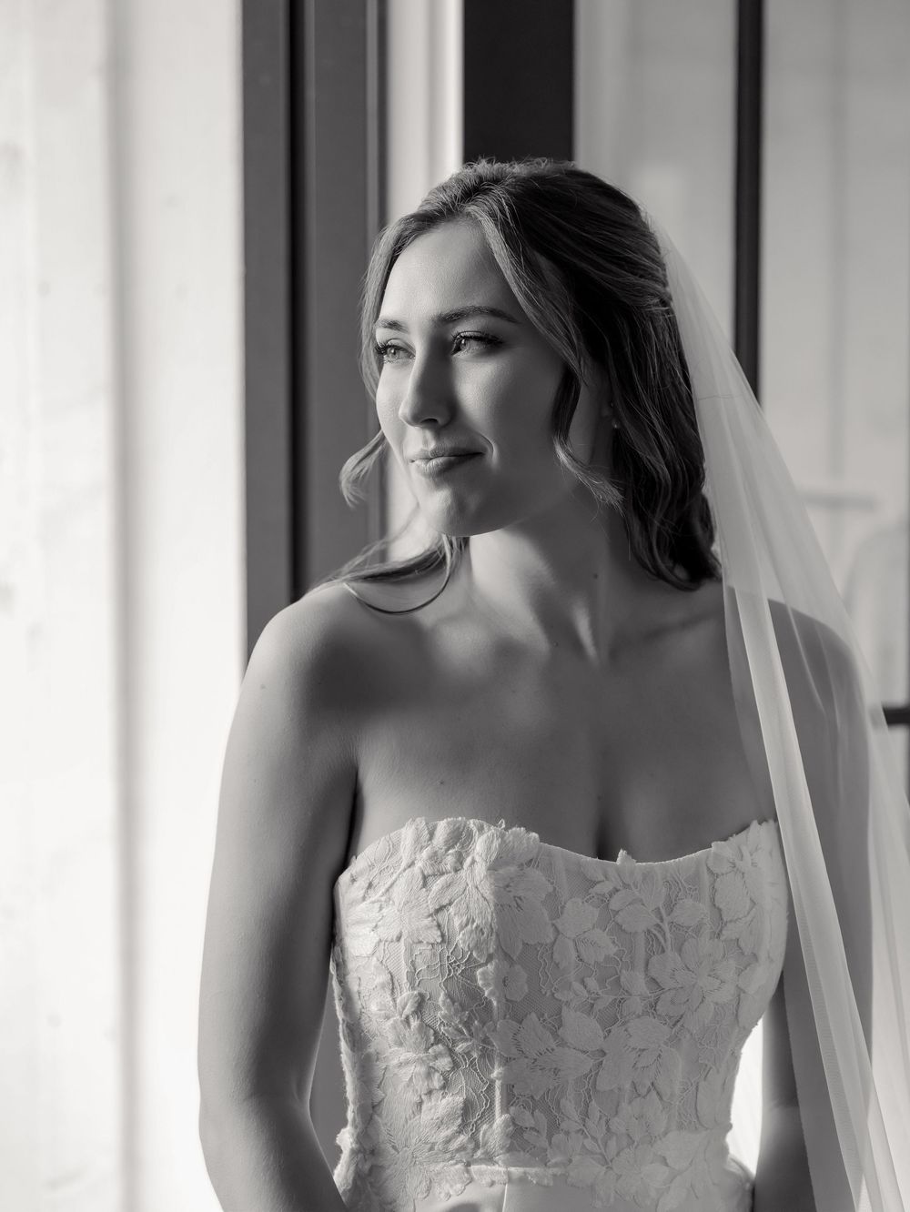 Black and white portrait of a bride in a strapless lace gown and veil gazing gracefully out a window.