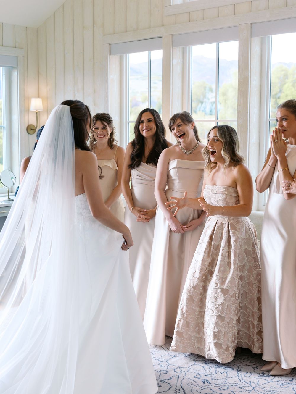 Bride reveals wedding dress to excited bridesmaids in blush gowns inside bright venue room.