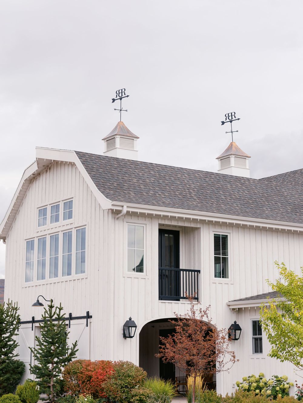 Elegant white barn with weather vanes, cupolas, and black accents surrounded by lush landscaping.