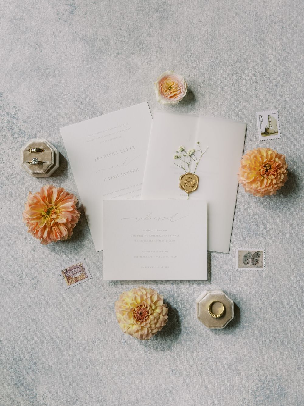 Elegant wedding invitation suite with gold wax seal, peach dahlias, vintage stamps, and ring boxes on gray background.