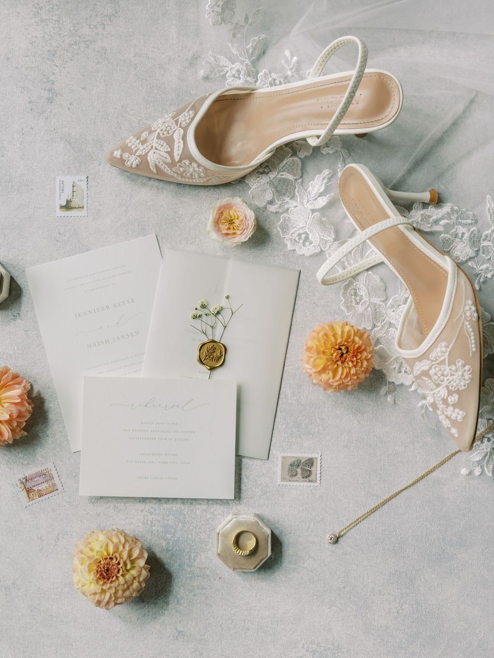 Elegant wedding flat lay featuring lace bridal heels, floral invitations with wax seal, orange dahlias, and a velvet ring box.