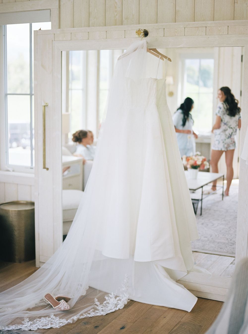Elegant white wedding gown with lace veil hanging in bright bridal suite as bridesmaids prepare in background.