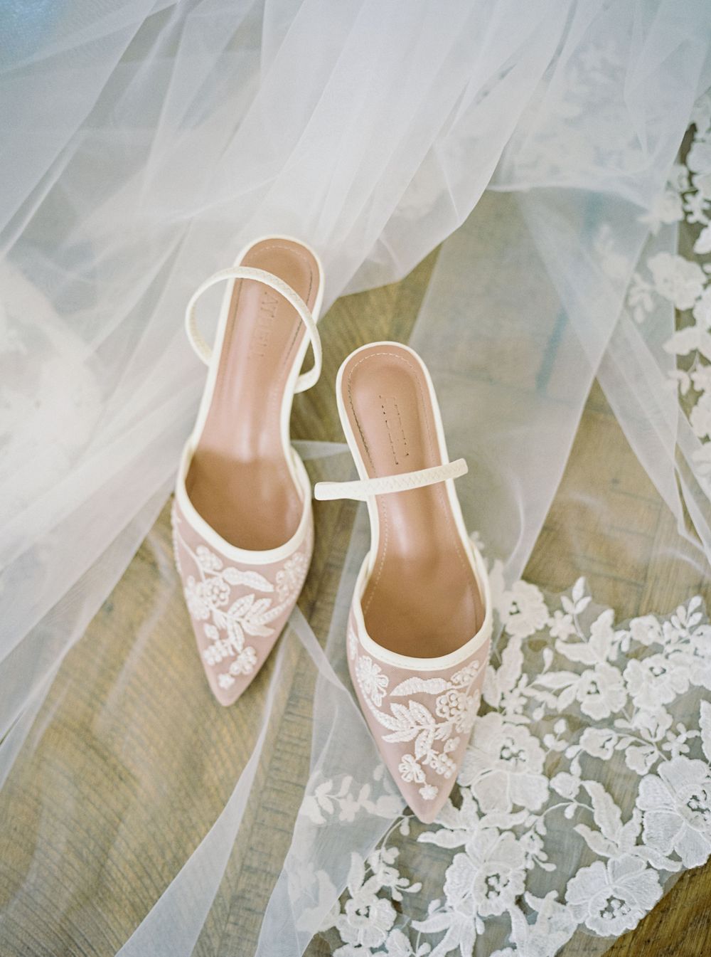 Elegant blush lace pointed-toe bridal mules with ivory ankle strap resting on a delicate lace wedding veil.