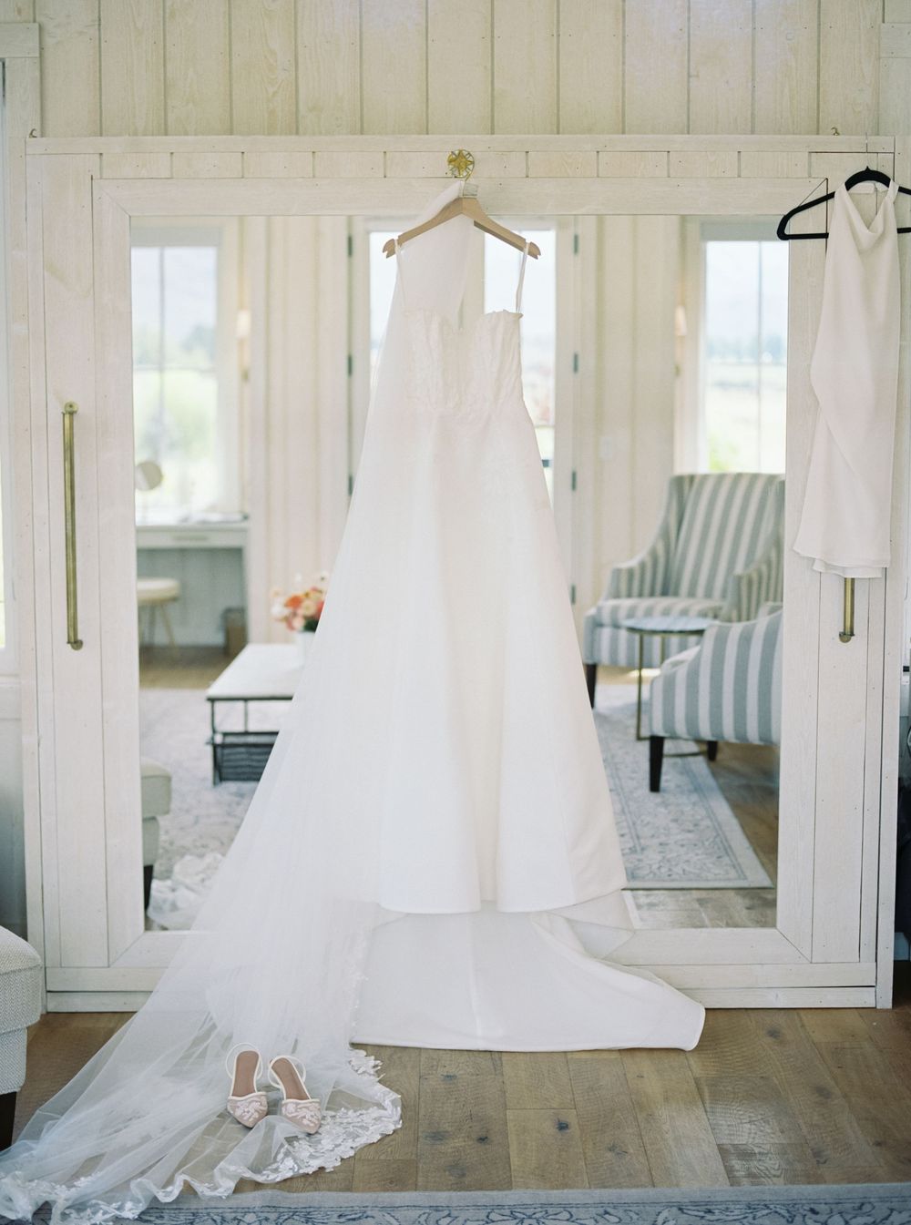 Elegant white A-line wedding gown hangs on mirrored doors above lace bridal heels in a bright, airy bridal suite.