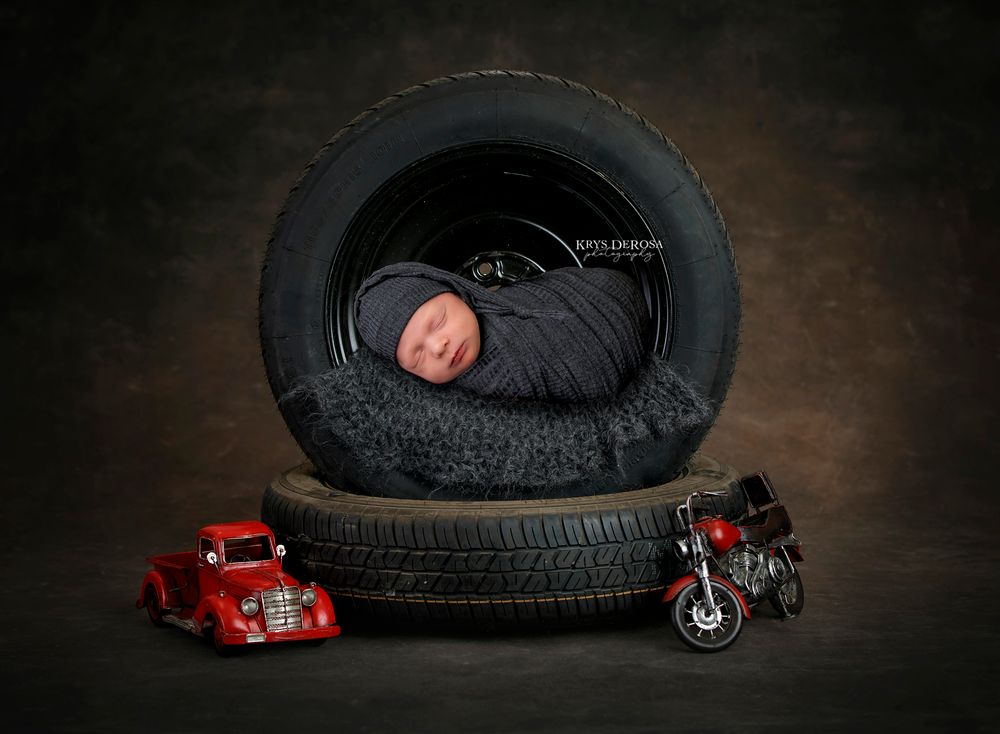 Newborn baby sleeping in a tire with toy red car and motorcycle, professional newborn photography studio portrait.