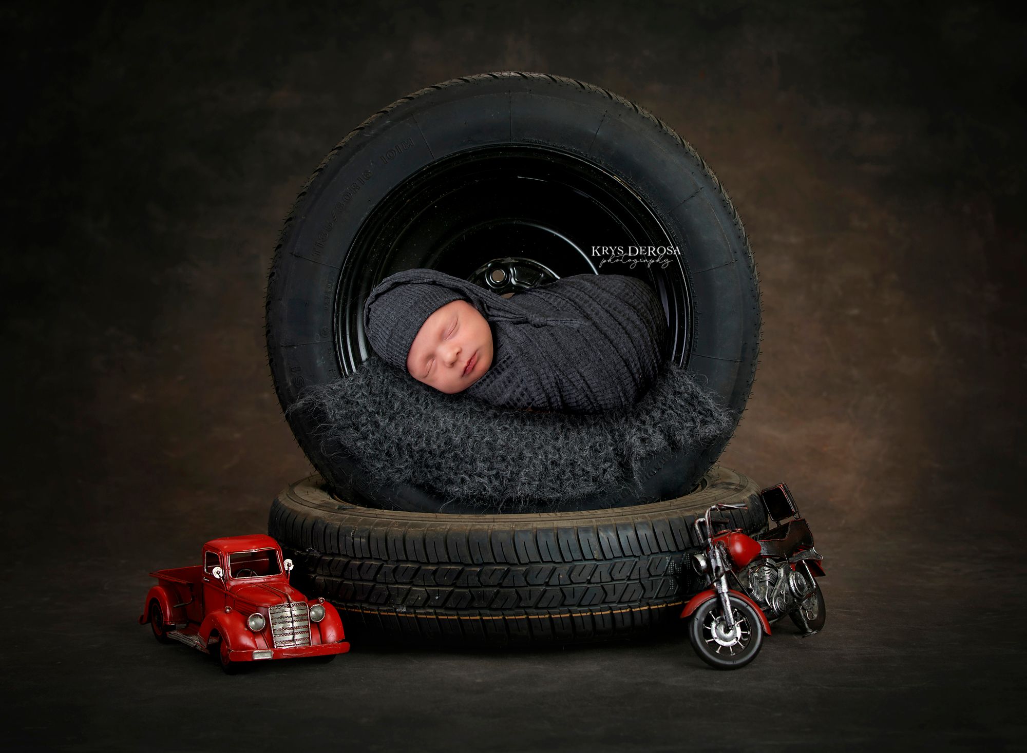 Newborn baby sleeping in a tire with toy red car and motorcycle, professional newborn photography studio portrait.