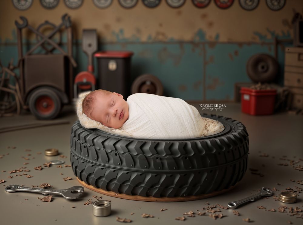 Newborn baby swaddled in white wrap sleeping peacefully inside a large tire in a mechanic garage setting.