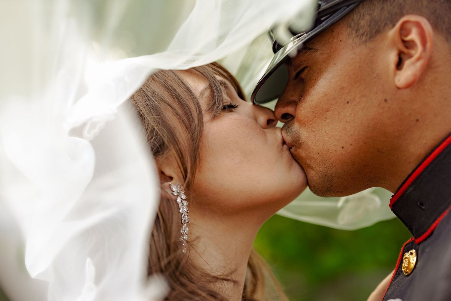 Bride and Marine groom share a romantic kiss under a flowing white veil outdoors.