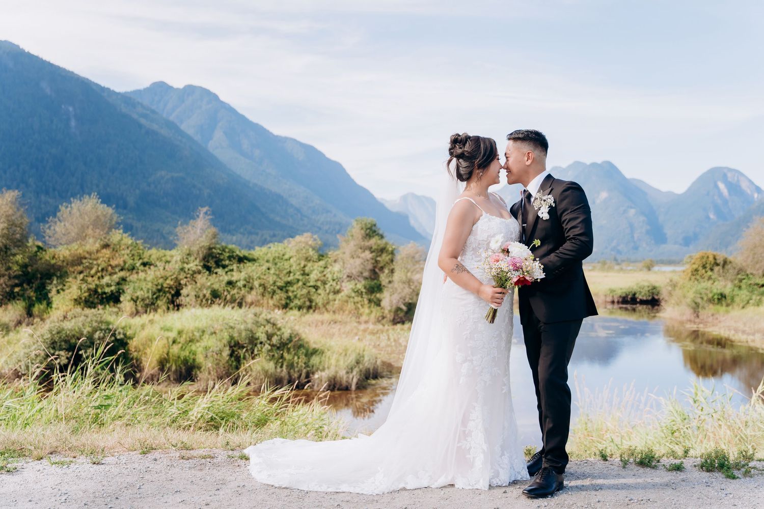 Bride and groom stand on rocky mountain summit overlooking snow-capped peaks and serene blue lake below.