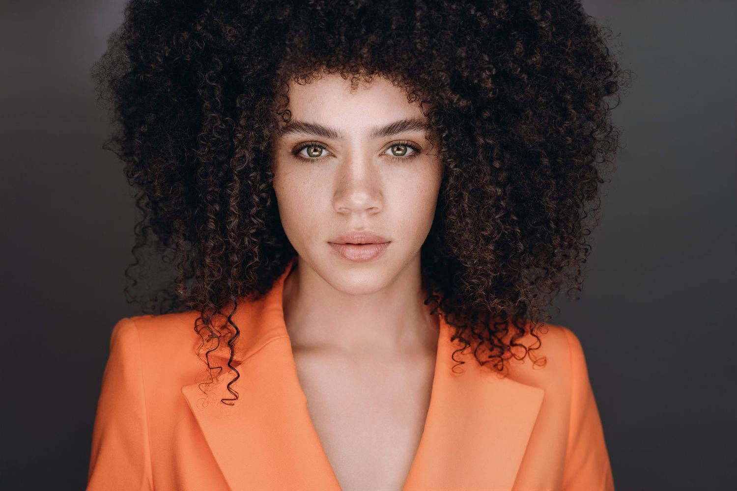 Young woman with large curly afro hair wearing orange blazer against gray background.