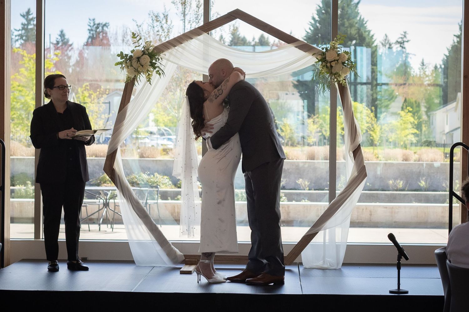 Bride and groom share first kiss under geometric hexagonal wooden arch with floral accents indoors.
