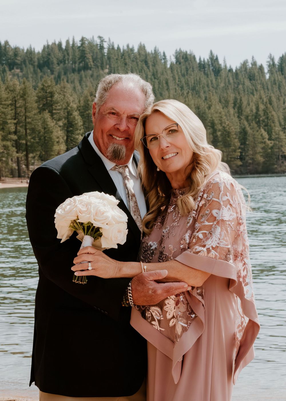 Couple in formal attire poses with white bouquet by a lake surrounded by pine trees, likely at a wedding ceremony.