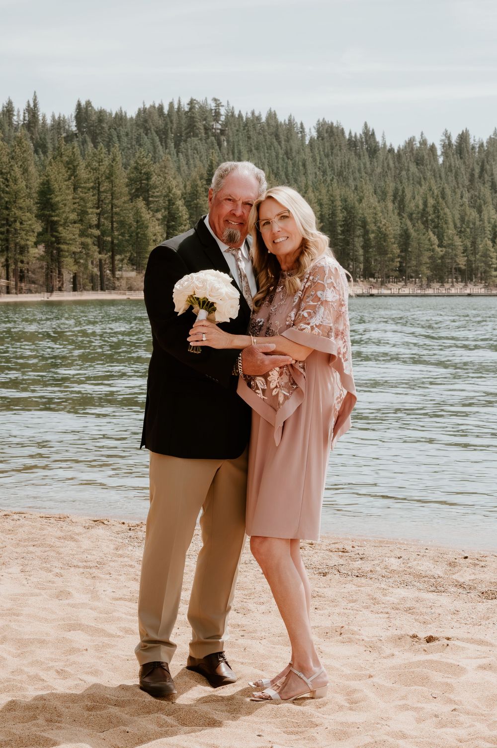 Couple posing with white bouquet on sandy lake shore with pine forest backdrop.