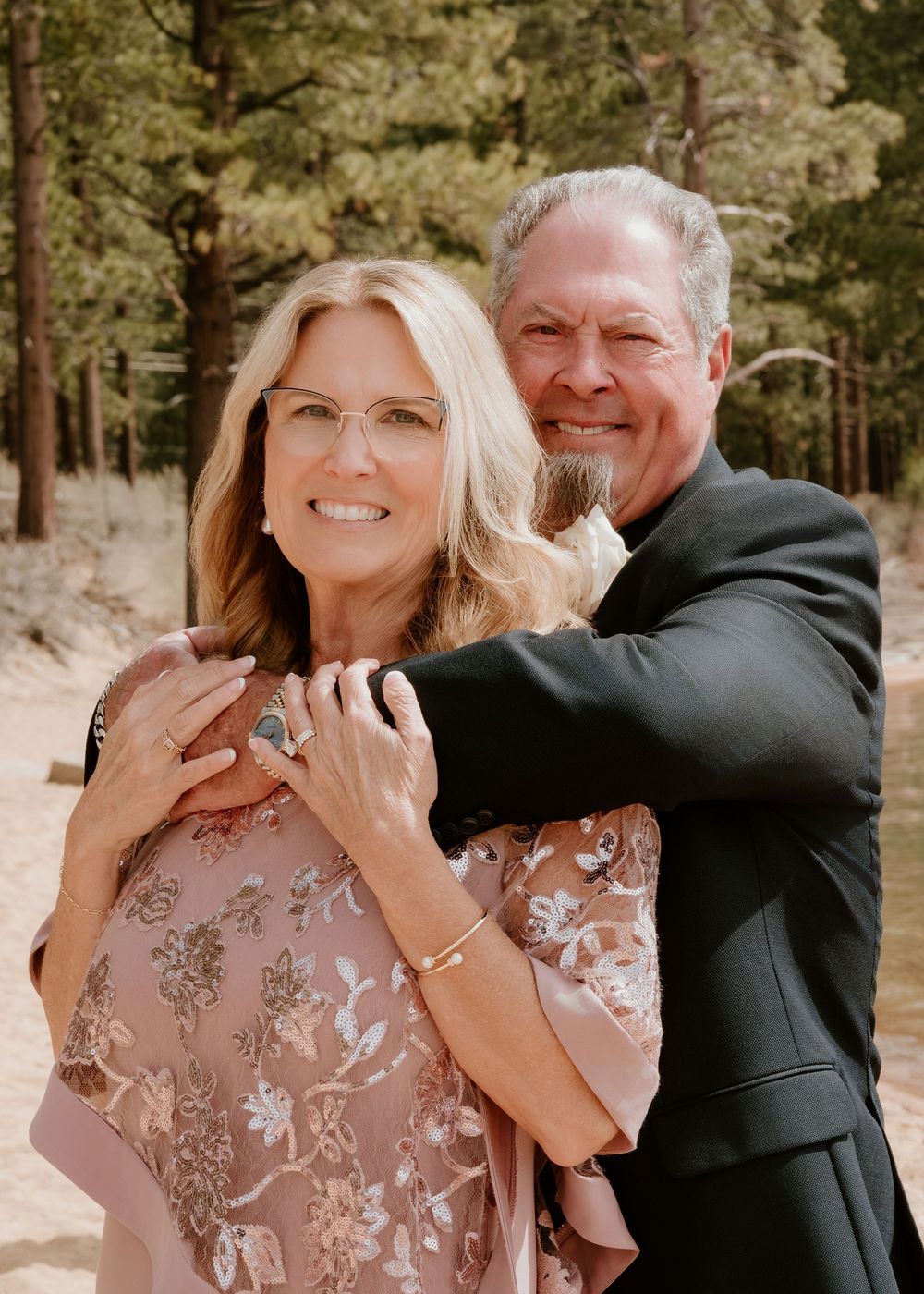 A smiling couple poses outdoors among pine trees, woman in floral pink dress, man in dark suit embracing her from behind.