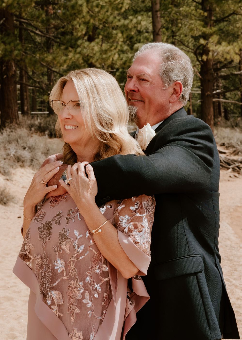 Couple in formal attire embracing outdoors among pine trees, woman in floral pink dress, man in black suit.