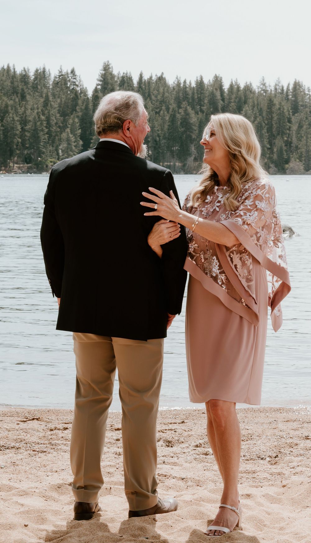 Elegant older couple standing on a sandy lakeside beach, woman in pink floral dress smiling at man in black blazer.