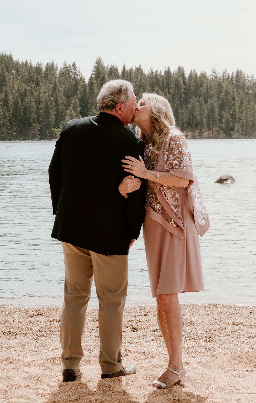 Couple sharing a romantic kiss on a sandy lakeside beach surrounded by pine trees.