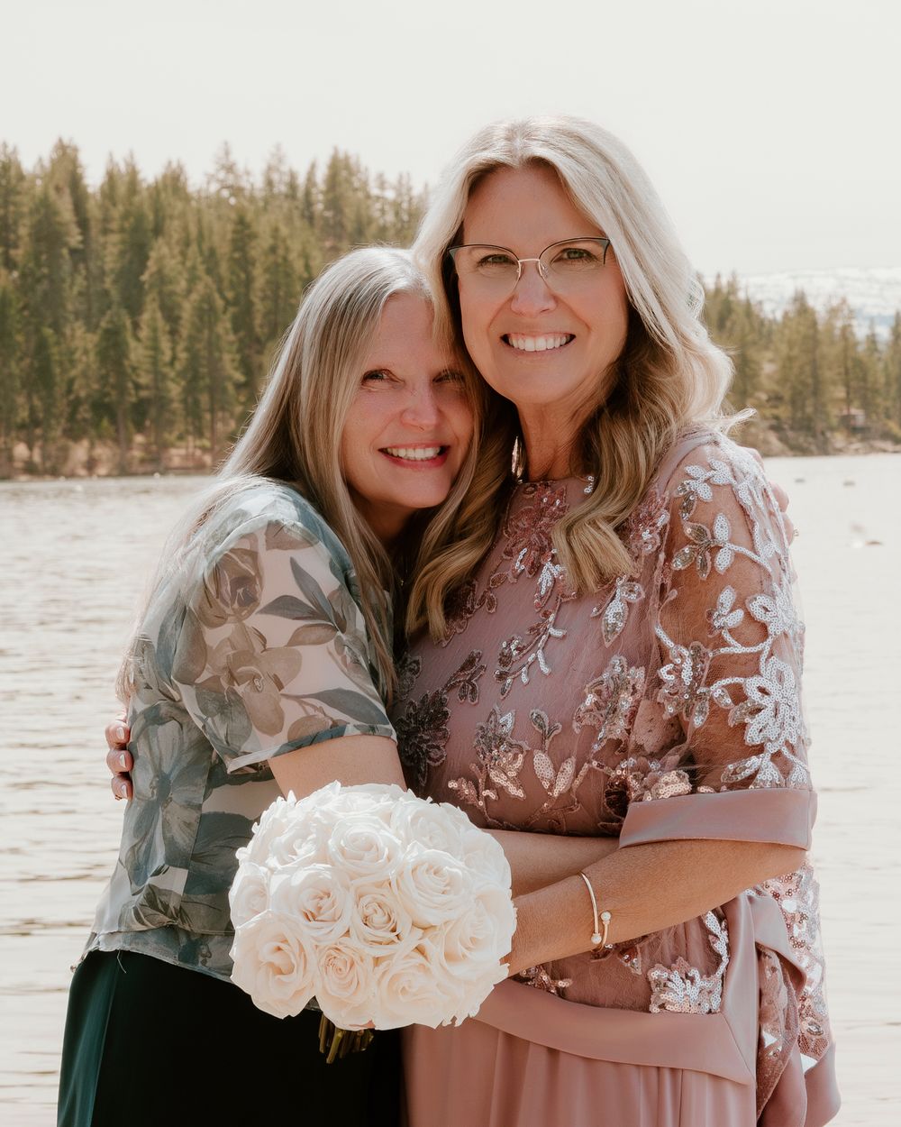 Two women smiling and embracing outdoors near a lake, one holding white roses bouquet, wearing elegant dresses.
