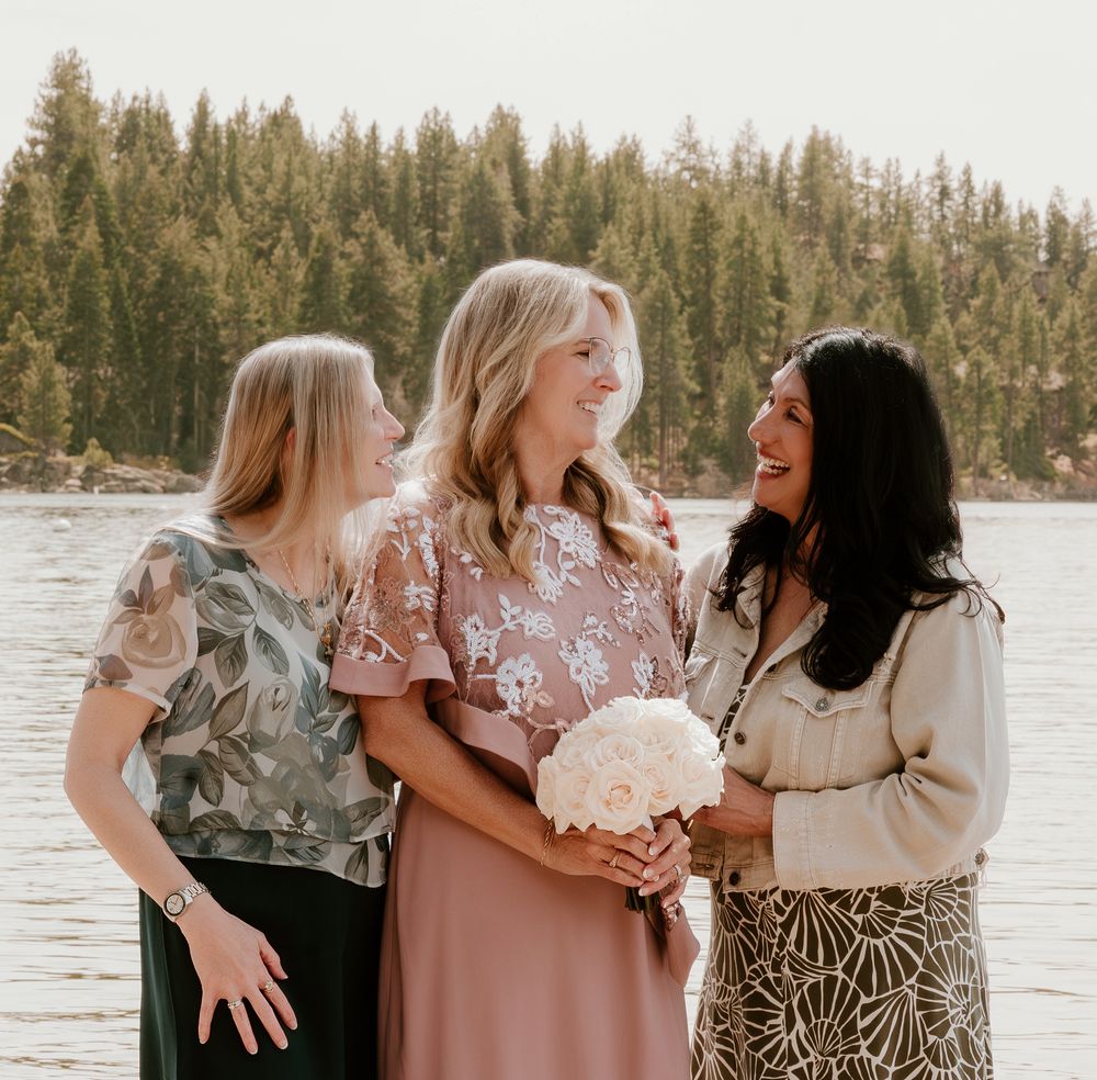 Three women smiling together outdoors near a lake, one holding a white bouquet, with pine trees in the background.