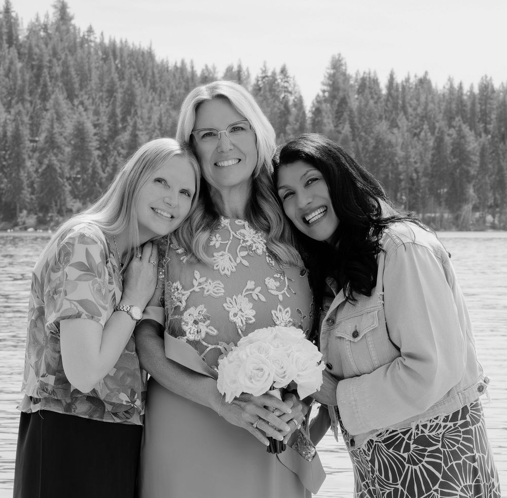 Three women smile together outdoors near a lake, with the center woman holding a white bouquet.