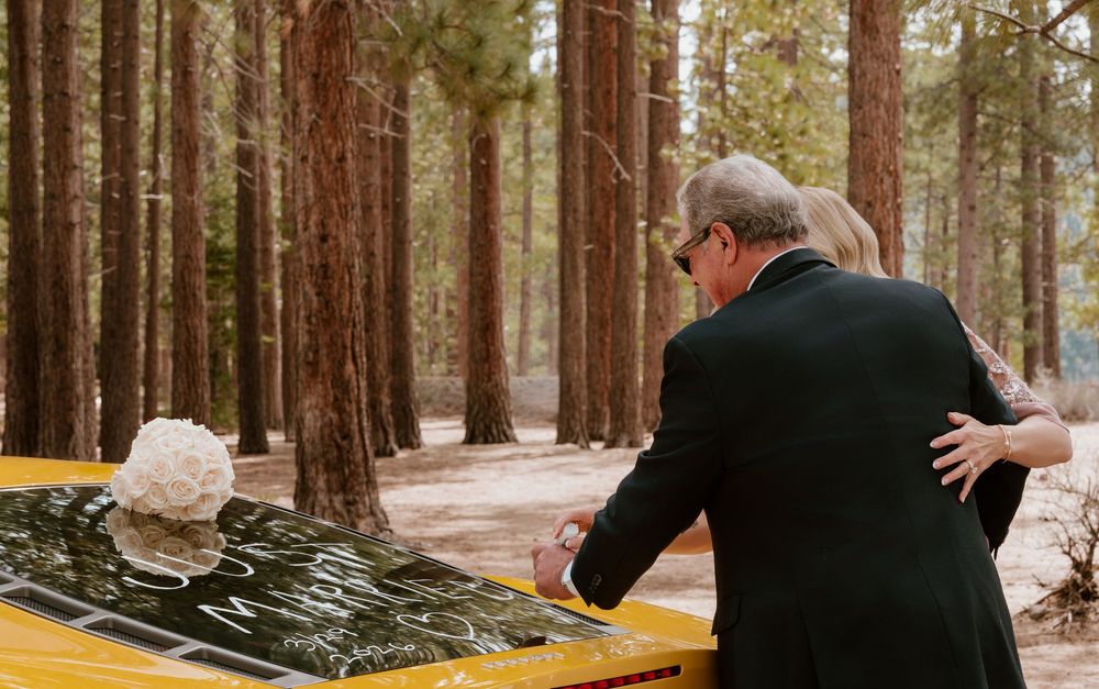 Couple signing wedding guest book on yellow car hood in pine forest, bride holding white bouquet.