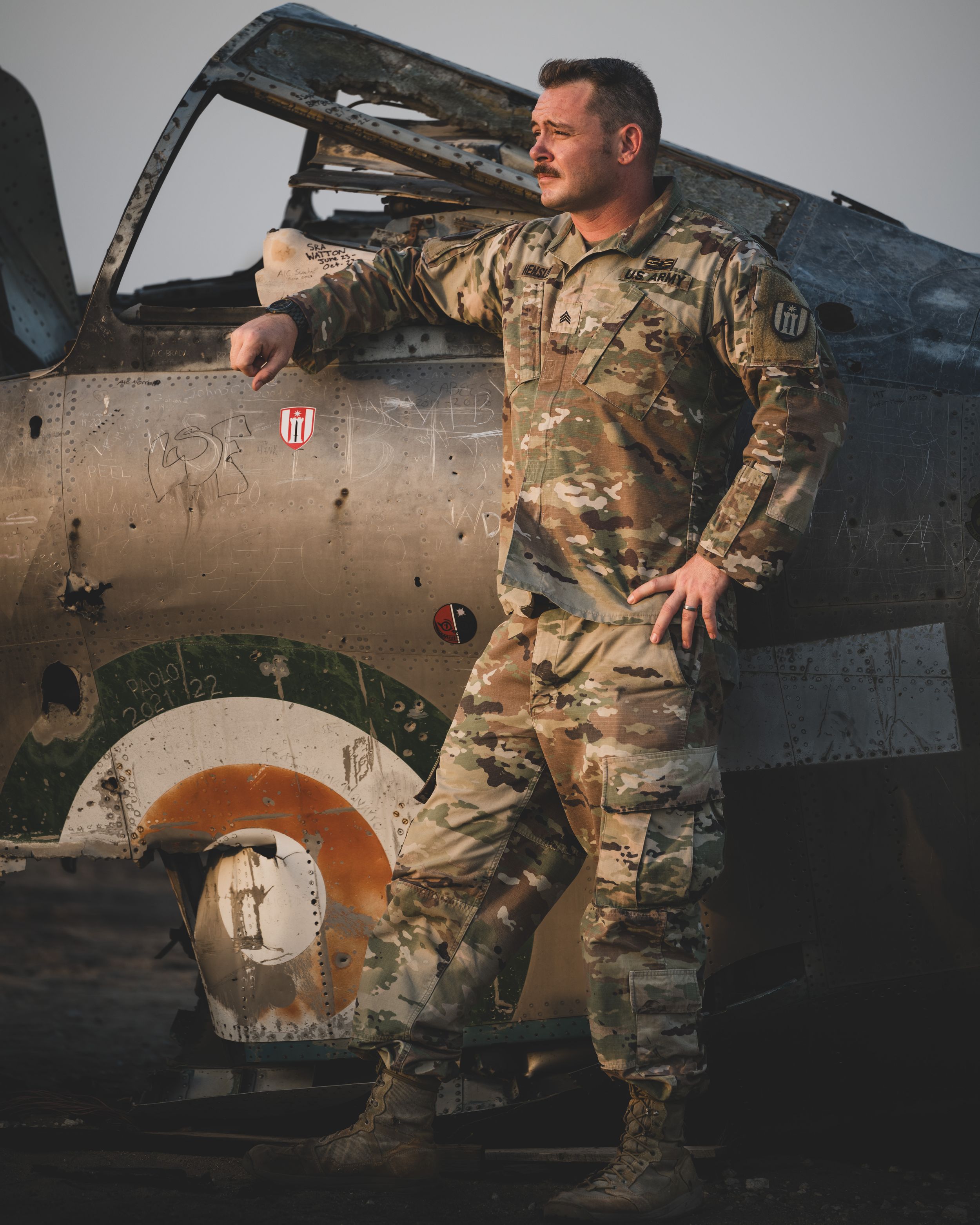 A soldier in camouflage uniform stands confidently beside a weathered military aircraft outdoors.