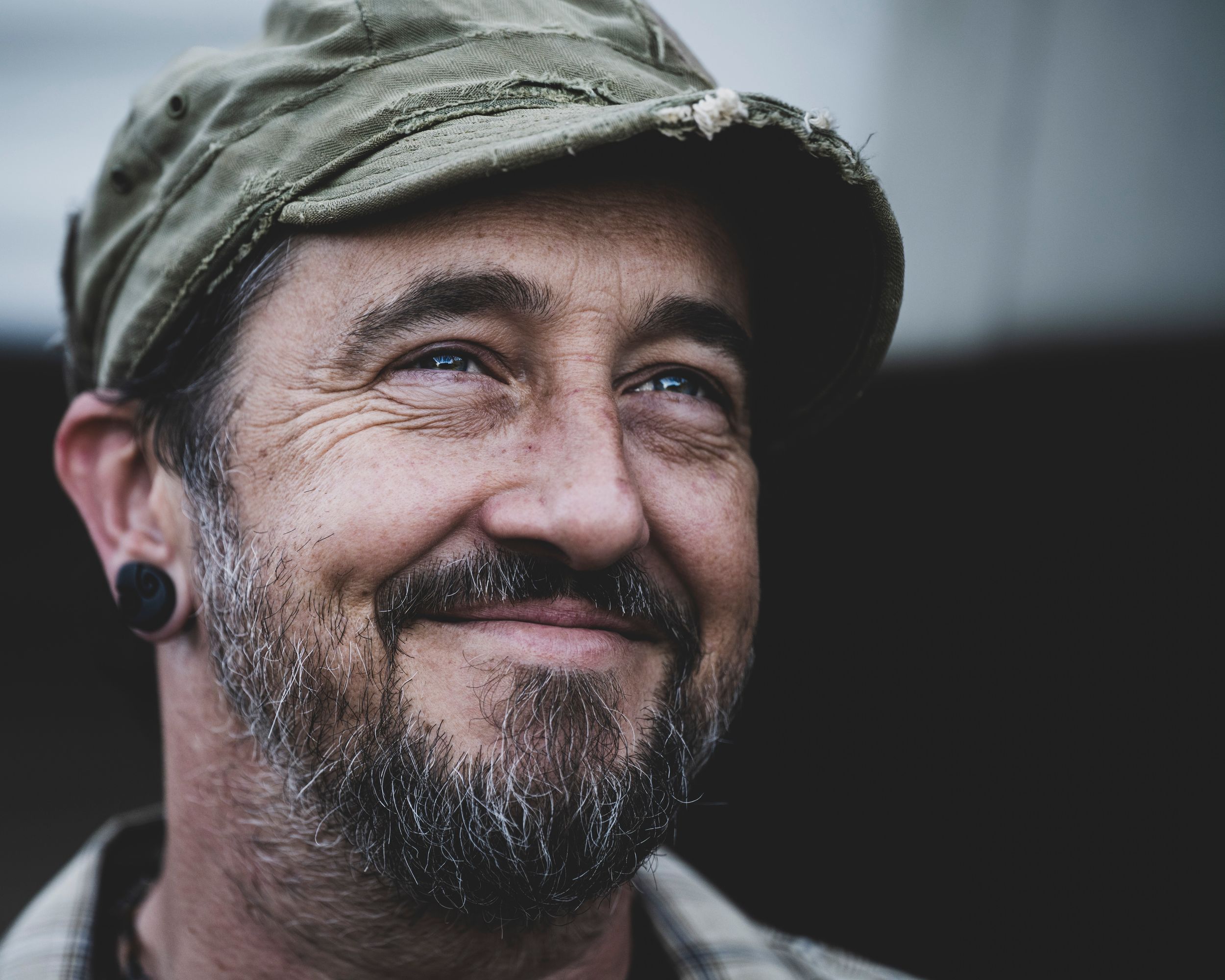 Smiling bearded man wearing a green cap and ear gauge, with blue eyes and a goatee, captured in a candid outdoor portrait.