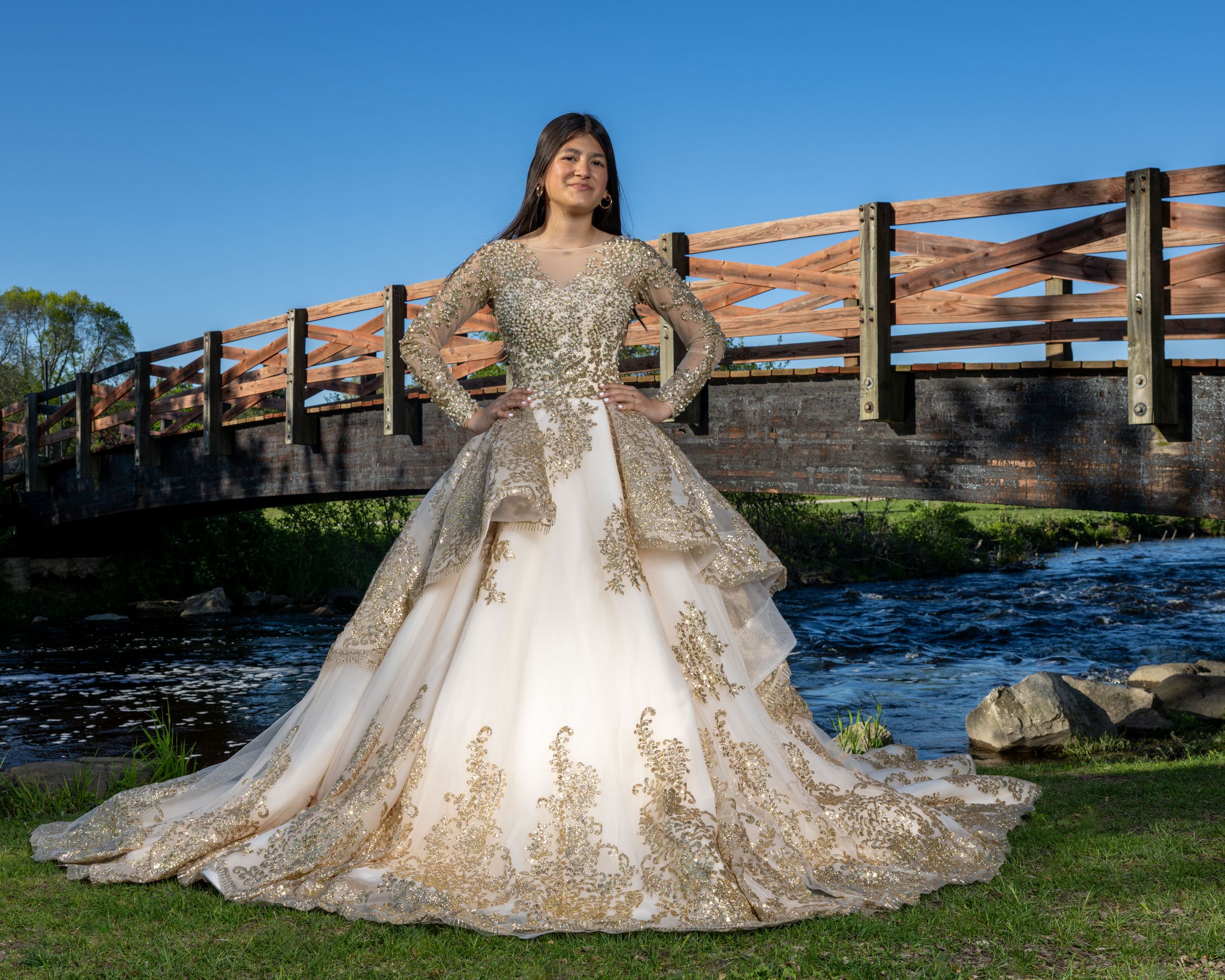 Young woman in elegant white and gold ball gown with long sleeves poses near a wooden bridge over a stream.