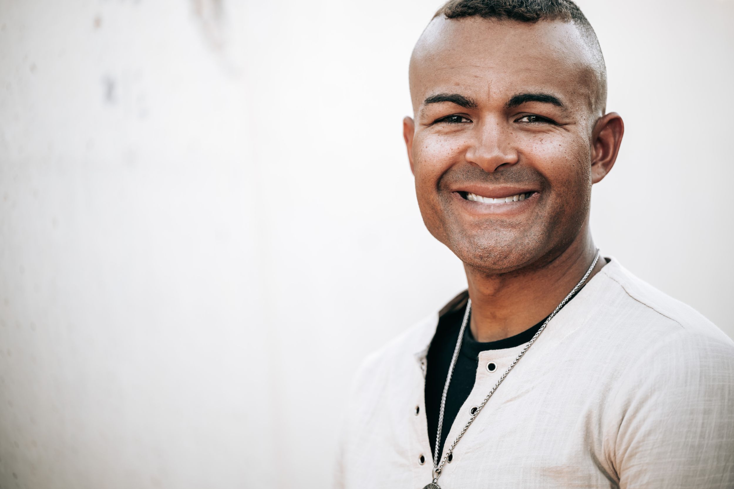 Smiling young Black man in a light beige shirt posing against a white wall in a confident portrait.