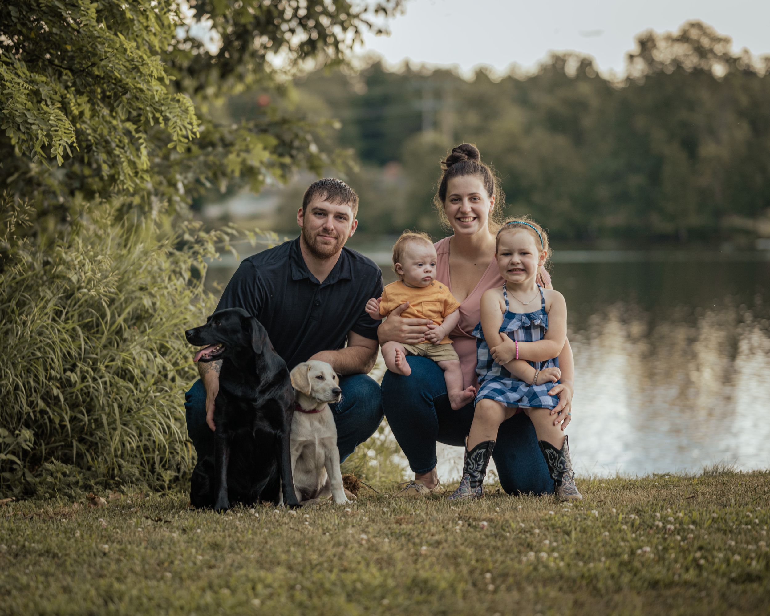 Family of four with two dogs posing by a serene lake surrounded by lush greenery for an outdoor portrait.