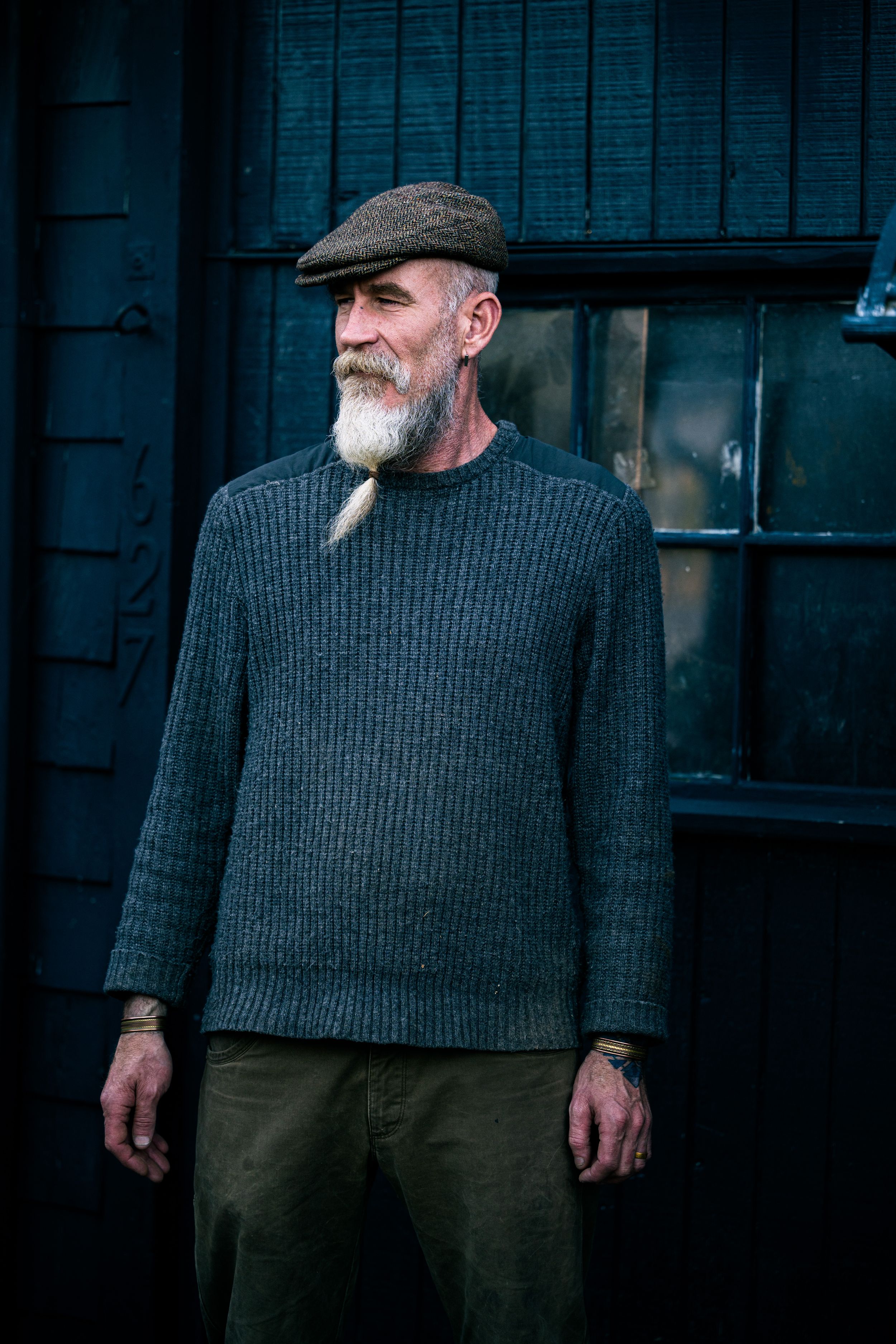 Older man with white beard wearing gray ribbed sweater and flat cap, standing outdoors near dark blue wooden wall.
