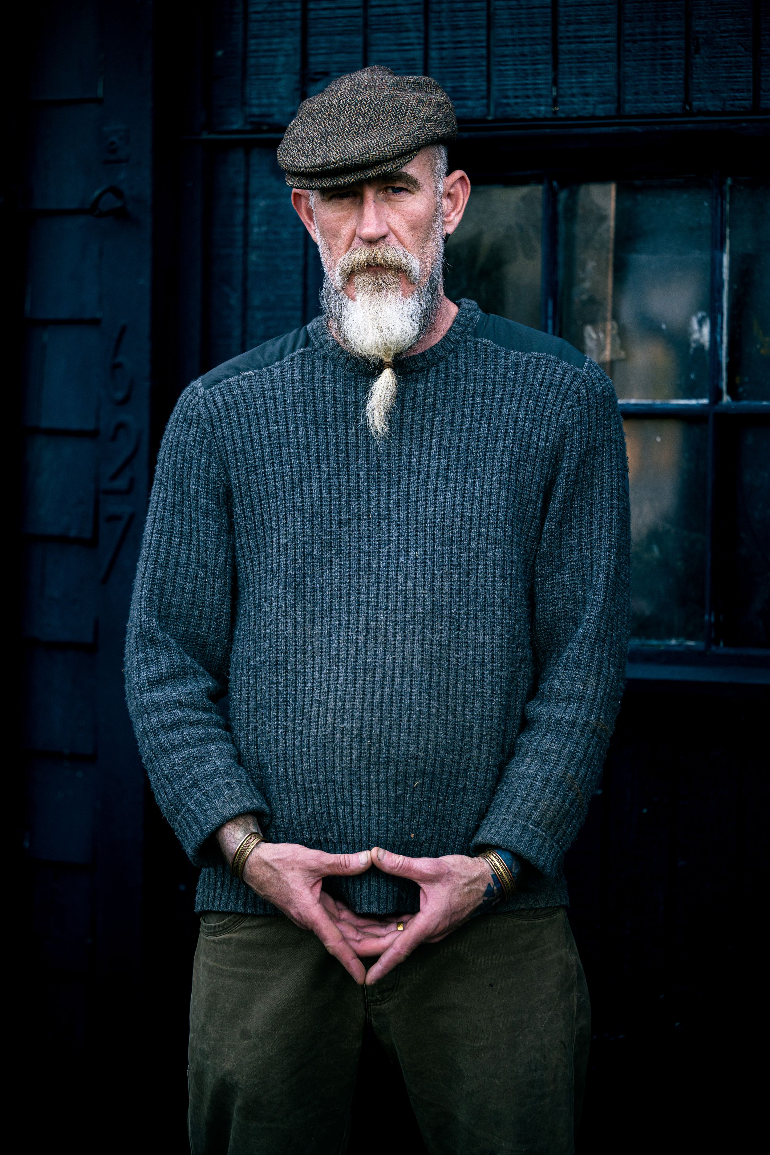 Older man with white beard wearing gray knit sweater and flat cap, standing with hands clasped outdoors.