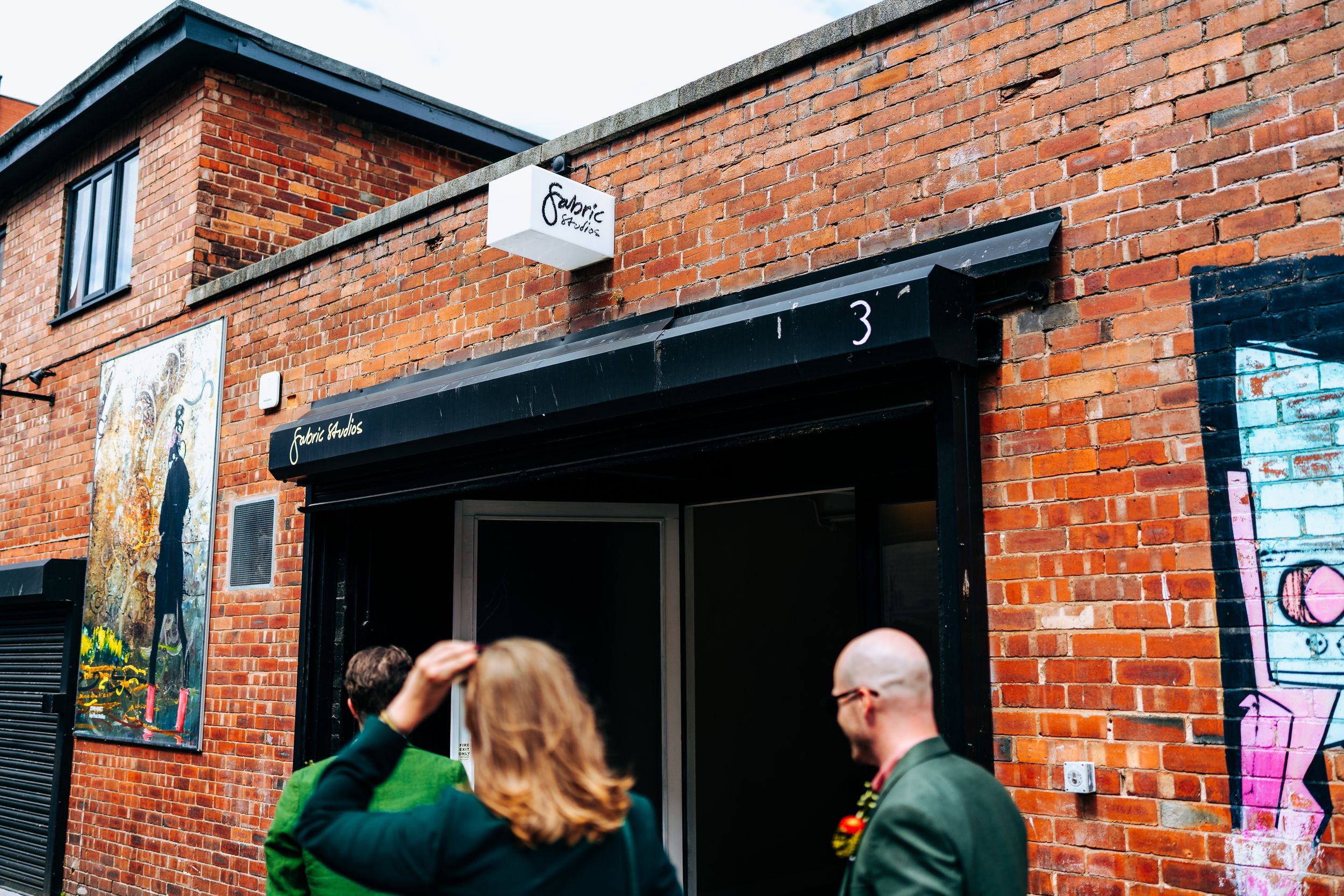 Wedding guests arriving at urban brick venue in Liverpool with street art, entering through black canopied entrance.