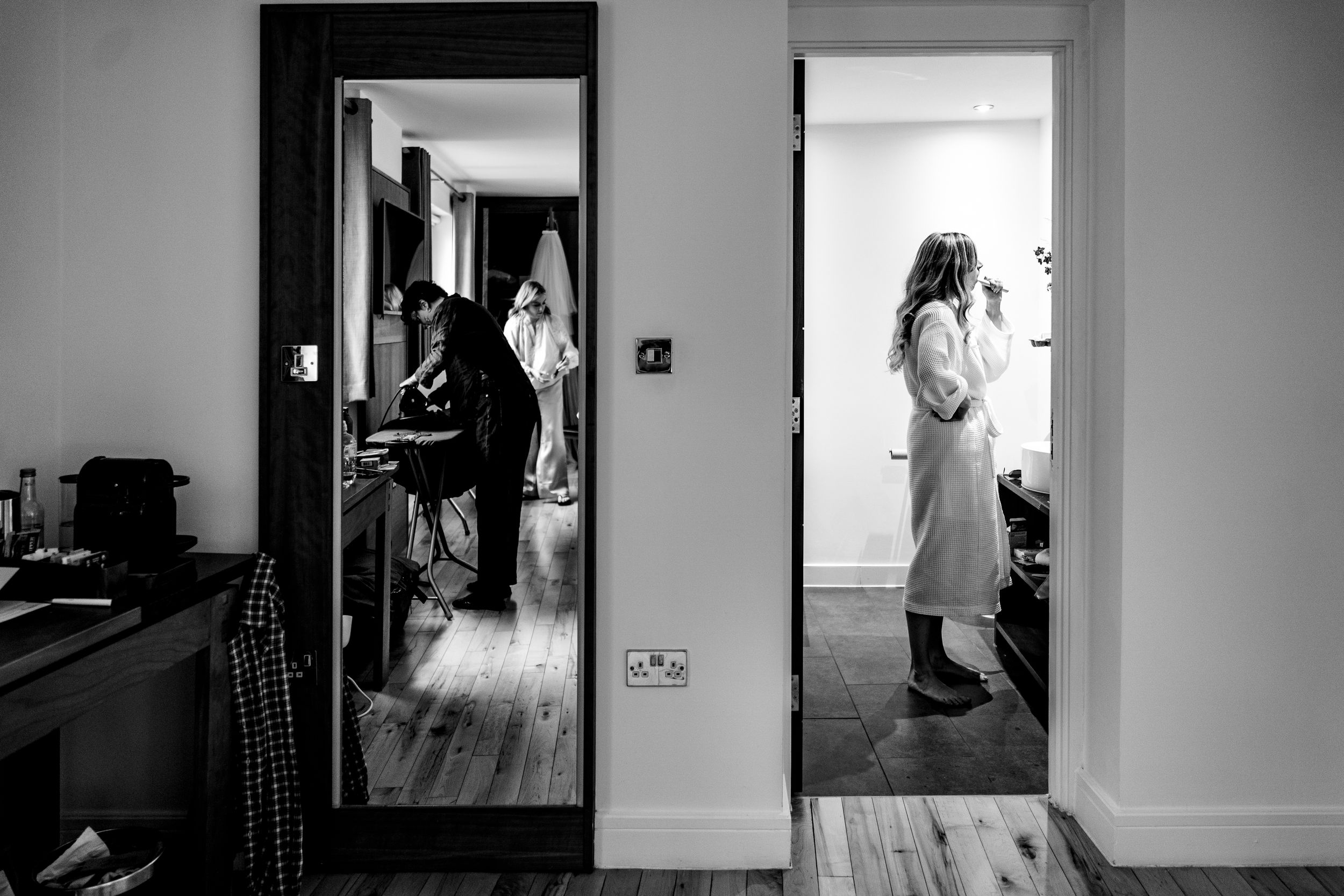 Black and white photo of bride getting ready in hotel room, reflected in mirror with bridesmaids nearby.