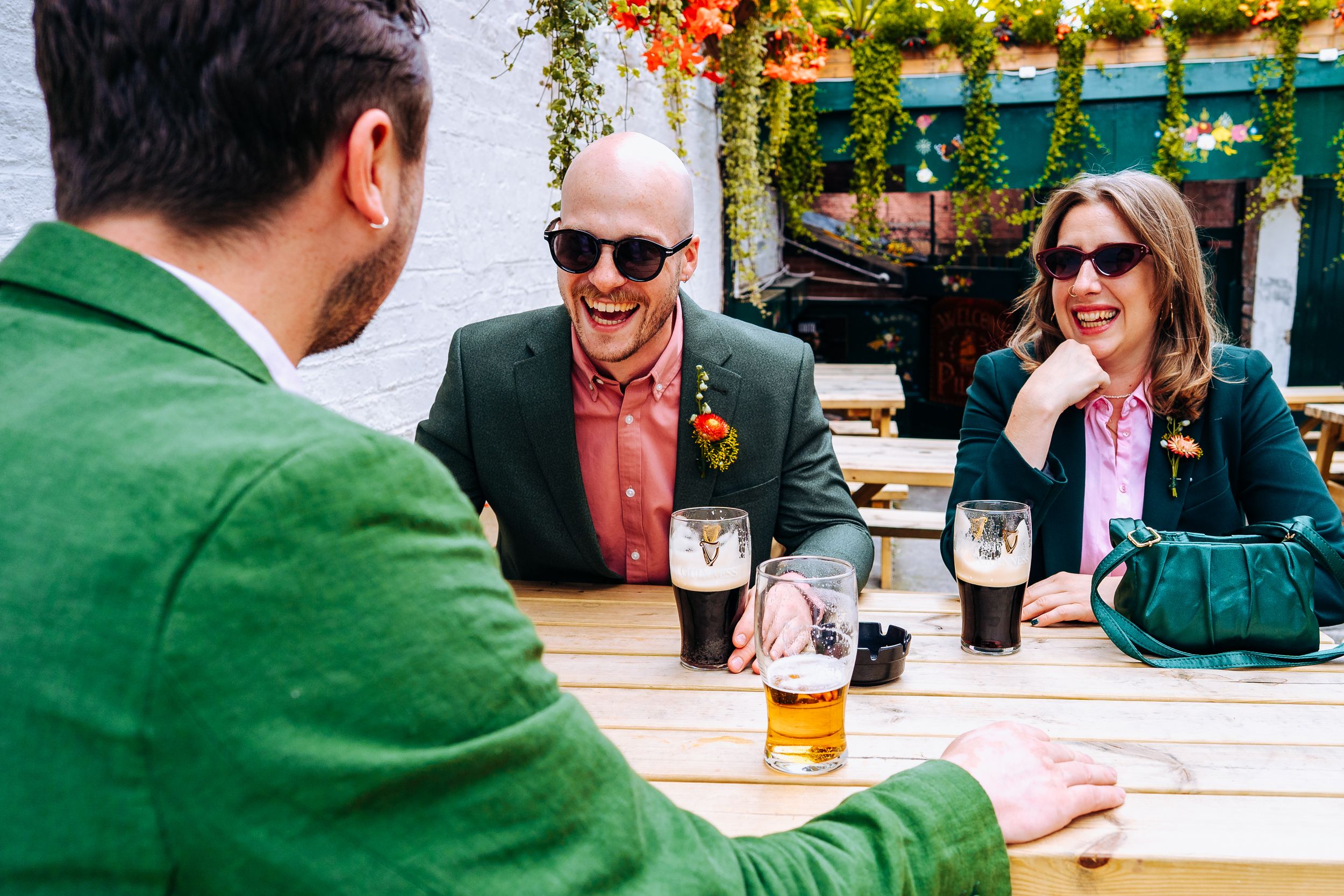 Wedding guests in colorful suits enjoying drinks at outdoor pub table on a sunny day in Liverpool.