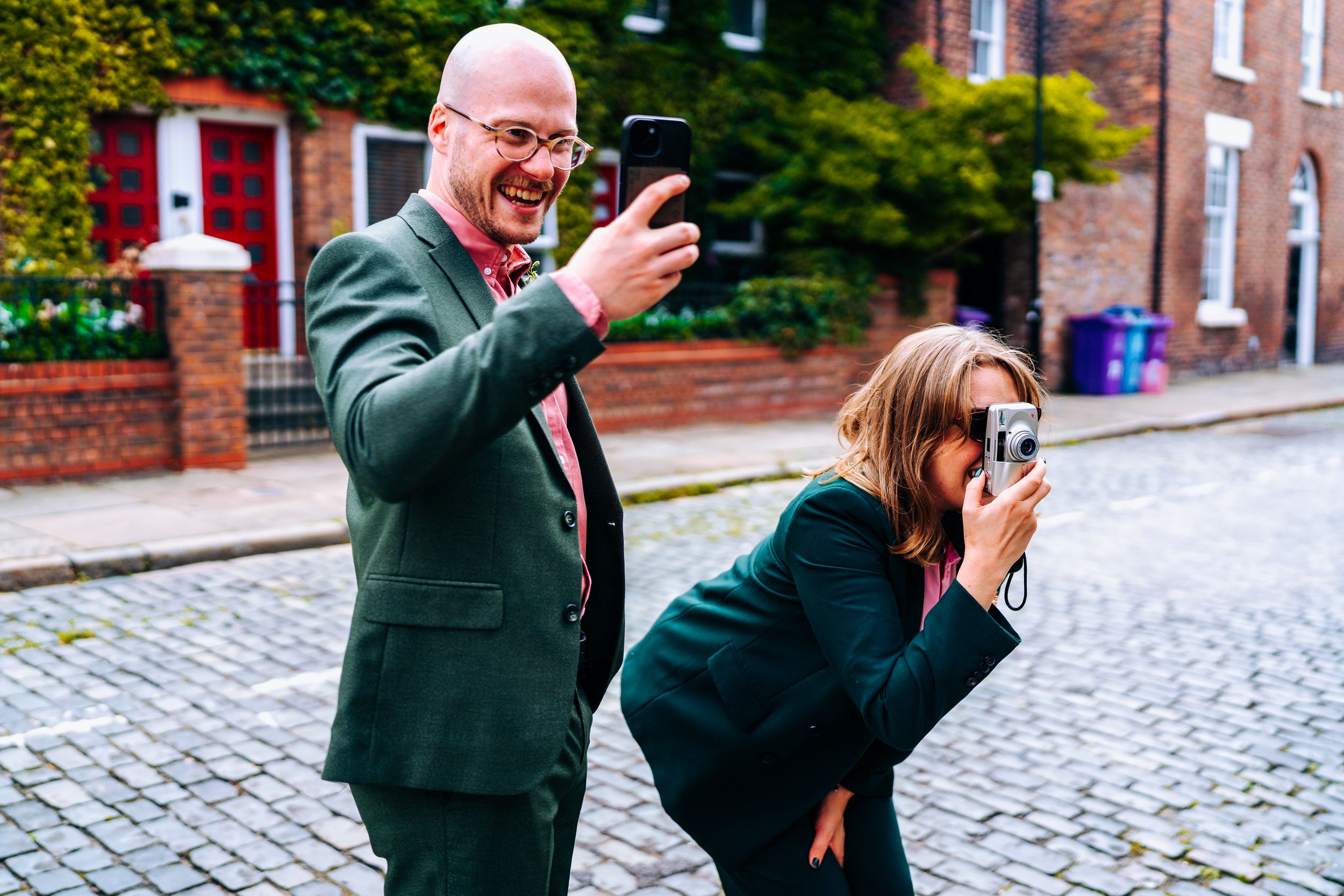 Two wedding guests photographing on a cobblestone street in Liverpool. Man with phone and woman with camera.