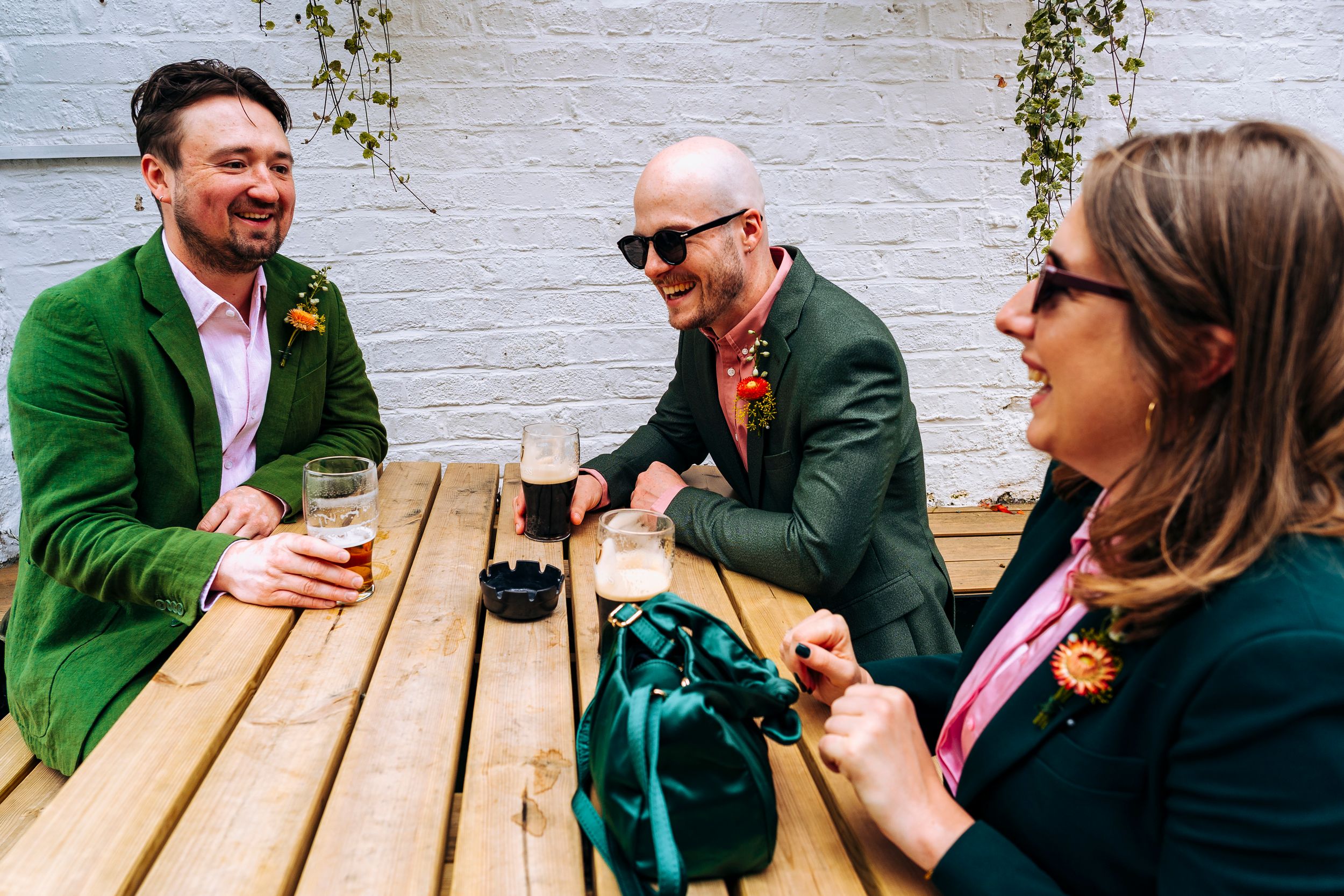 Two grooms in green suits laugh with a guest over drinks at an outdoor wooden table.