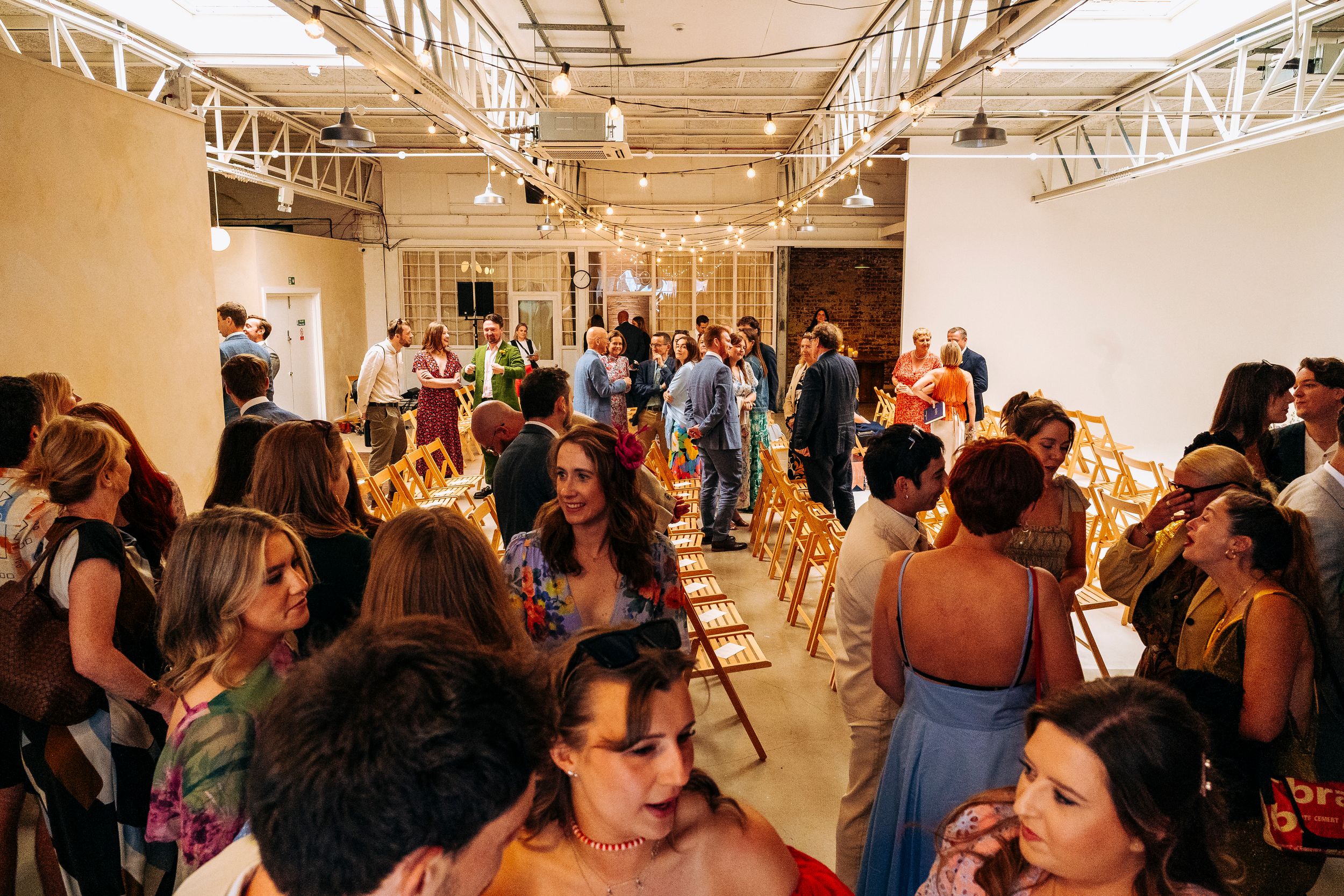 Wedding guests mingle in a bright industrial venue with wooden chairs.