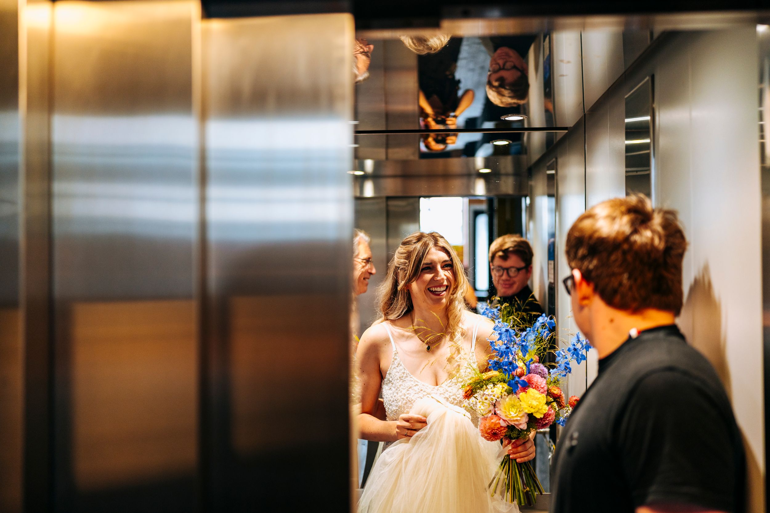 Smiling bride holding colorful bouquet steps out of elevator on wedding day.
